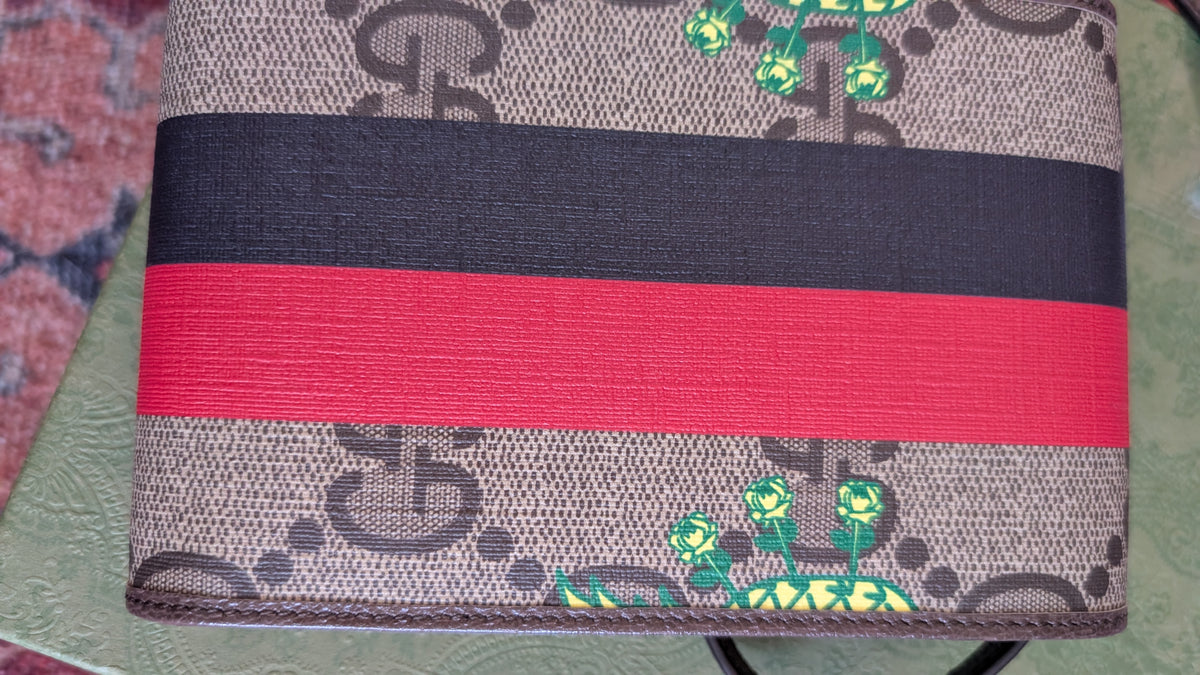 A close-up of a Gucci monogram canvas featuring the classic brown and black GG pattern, a wide black stripe, a red stripe, and green floral accents near the bottom. Pre-Owned Gucci x Ken Scott "What's Inside" Crossbody Phone Holder – GG Supreme Canvas – Full Set