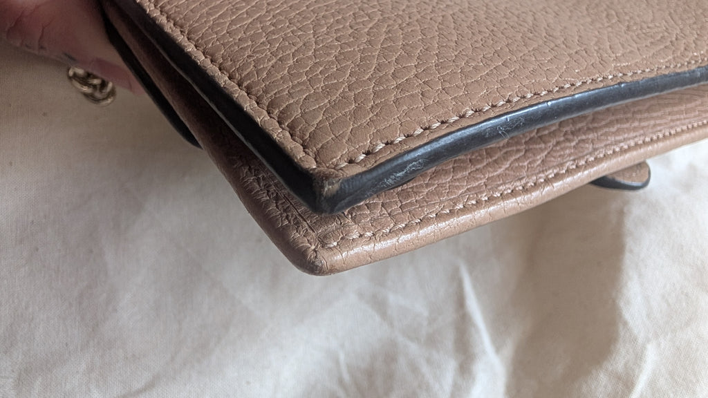 A close-up of a taupe pebbled leather clutch or wallet being held by a hand, showing the corner seam and black edge trim against a white fabric background.Pre-Owned Gucci Interlocking G Chain Wallet / Crossbody – Beige Pebbled Leather