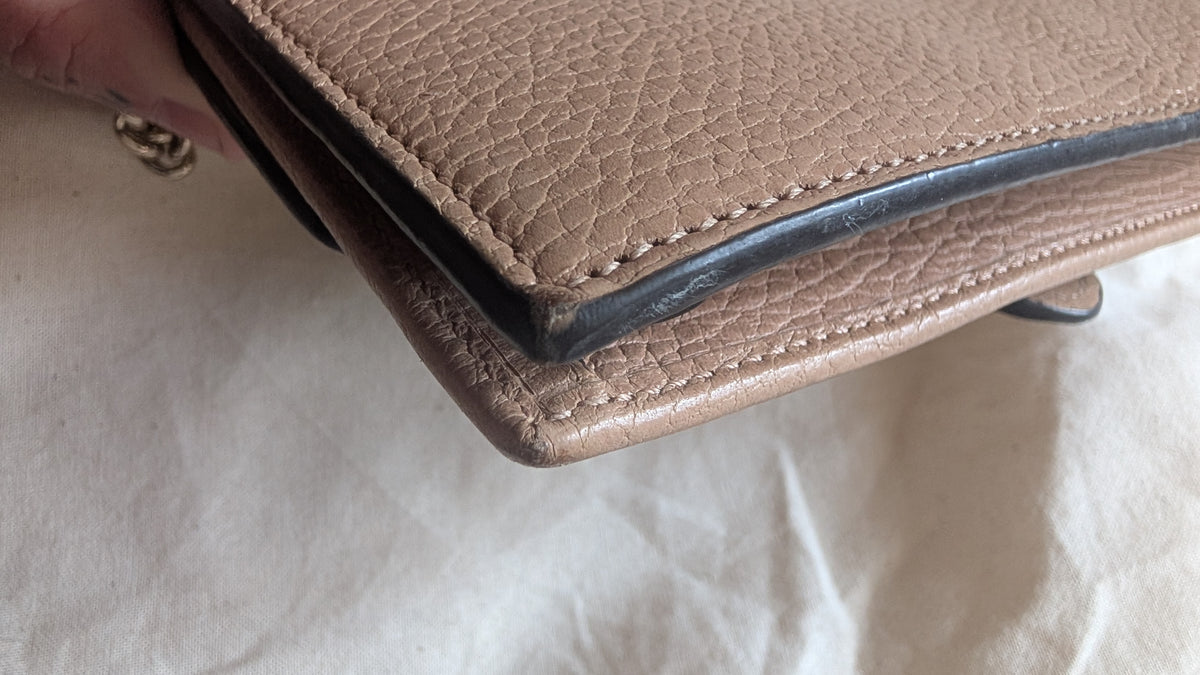 A close-up of a taupe pebbled leather clutch or wallet being held by a hand, showing the corner seam and black edge trim against a white fabric background.Pre-Owned Gucci Interlocking G Chain Wallet / Crossbody – Beige Pebbled Leather