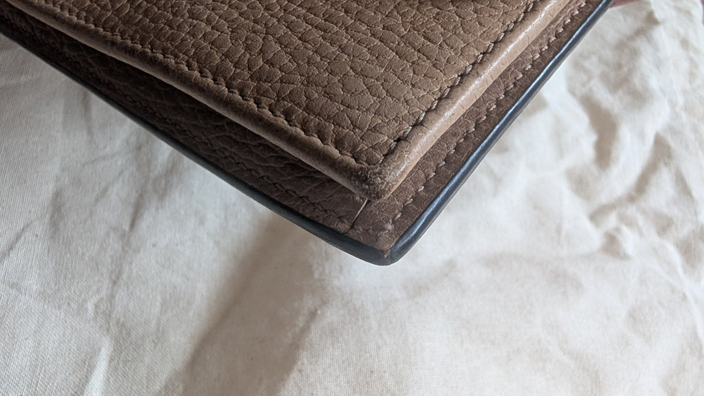 A close-up of a pebbled leather wallet corner with dark edging, resting on a textured off-white fabric background. Pre-Owned Gucci Interlocking G Chain Wallet / Crossbody – Beige Pebbled Leather