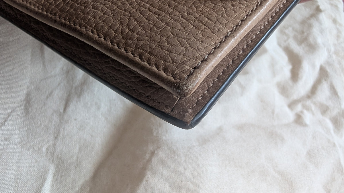 A close-up of a pebbled leather wallet corner with dark edging, resting on a textured off-white fabric background. Pre-Owned Gucci Interlocking G Chain Wallet / Crossbody – Beige Pebbled Leather