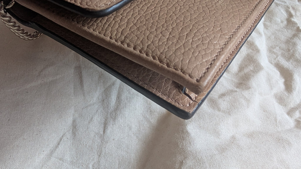 A close-up of a taupe pebbled leather handbag corner with black trim and a silver chain strap, resting on a textured off-white fabric surface. Pre-Owned Gucci Interlocking G Chain Wallet / Crossbody – Beige Pebbled Leather