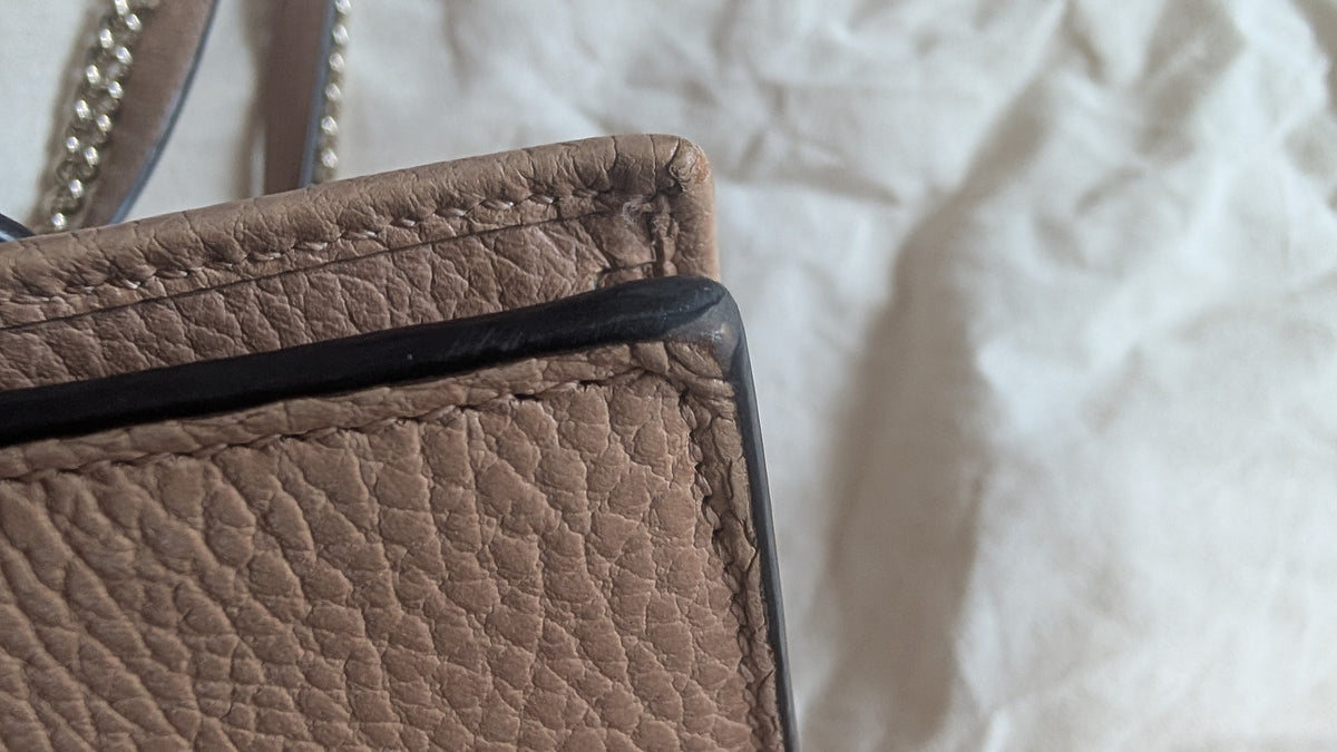 Close-up of a pebbled leather handbag corner with a black trim and silver chain strap, set against a wrinkled white fabric background. Pre-Owned Gucci Interlocking G Chain Wallet / Crossbody – Beige Pebbled Leather