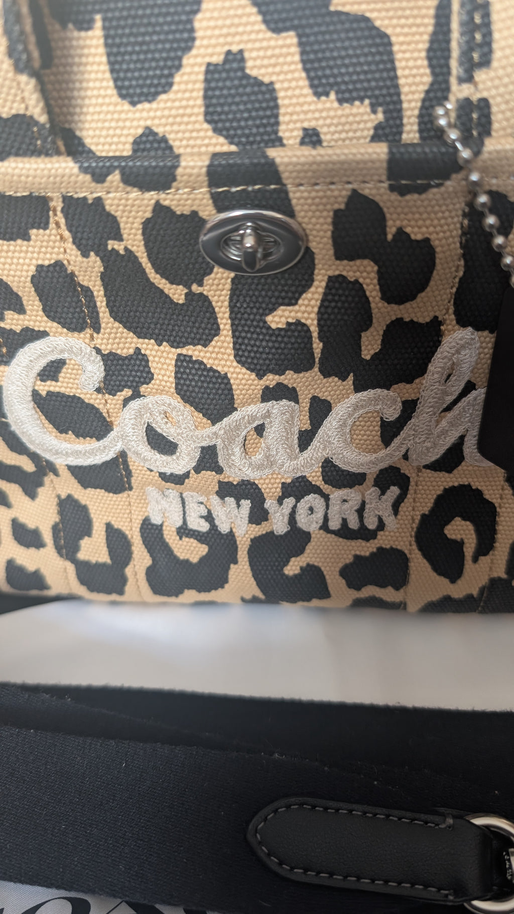 Close-up of a Coach New York leopard print handbag with a silver-tone metal snap button and embroidered Coach logo in silver on a beige and black patterned canvas. Pre-Owned Coach Cargo Tote  Leopard Print Canvas Bag