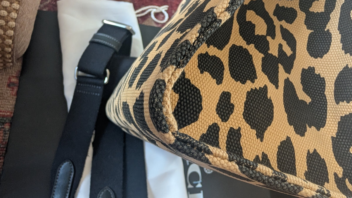 Close-up of a leopard print bag corner showing a tan and black patterned textile surface with black trim, alongside a black strap and white dust bag. Pre-Owned Coach Cargo Tote  Leopard Print Canvas Bag
