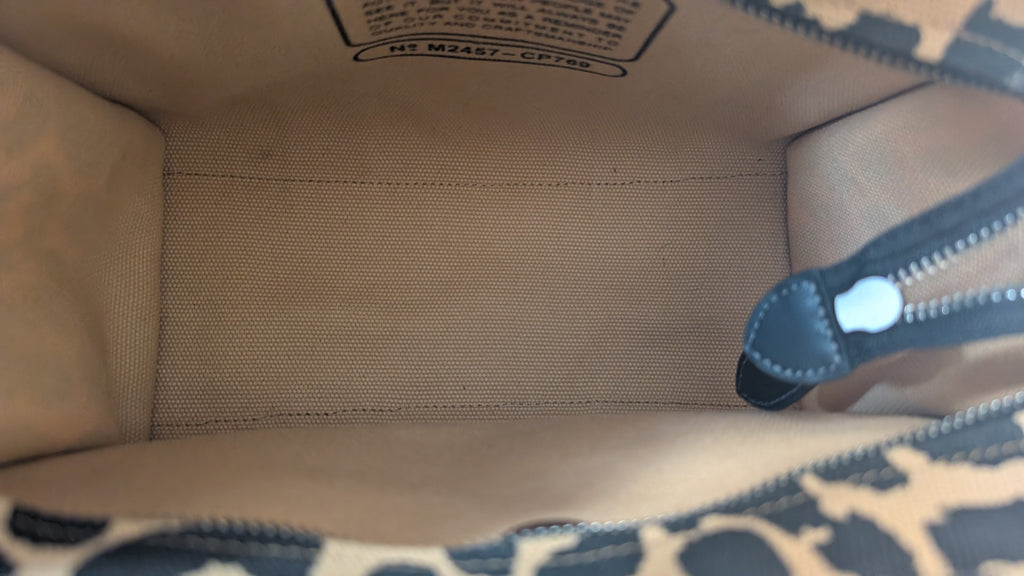 Close-up view of the interior of a handbag or tote, showing a beige woven fabric lining, a black zipper pull, and part of a dark patterned exterior at the bottom edge. Pre-Owned Coach Cargo Tote  Leopard Print Canvas Bag