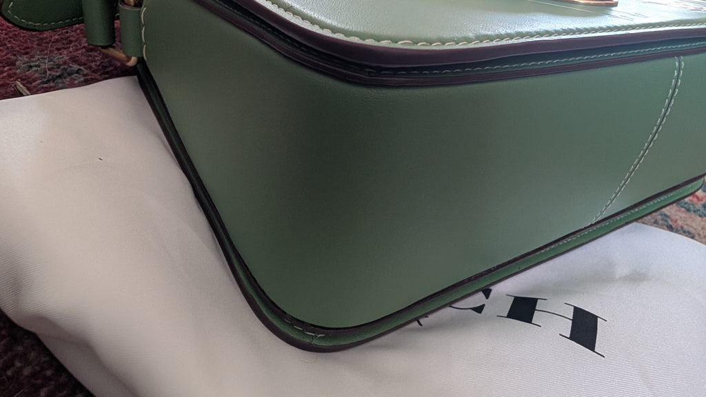A close-up of a  green leather handbag resting on a white dust bag with a black logo, set against a dark patterned carpet. Pre-Owned Coach x Disney Tabby Shoulder Bag – Mickey Mouse – Green – Limited Edition