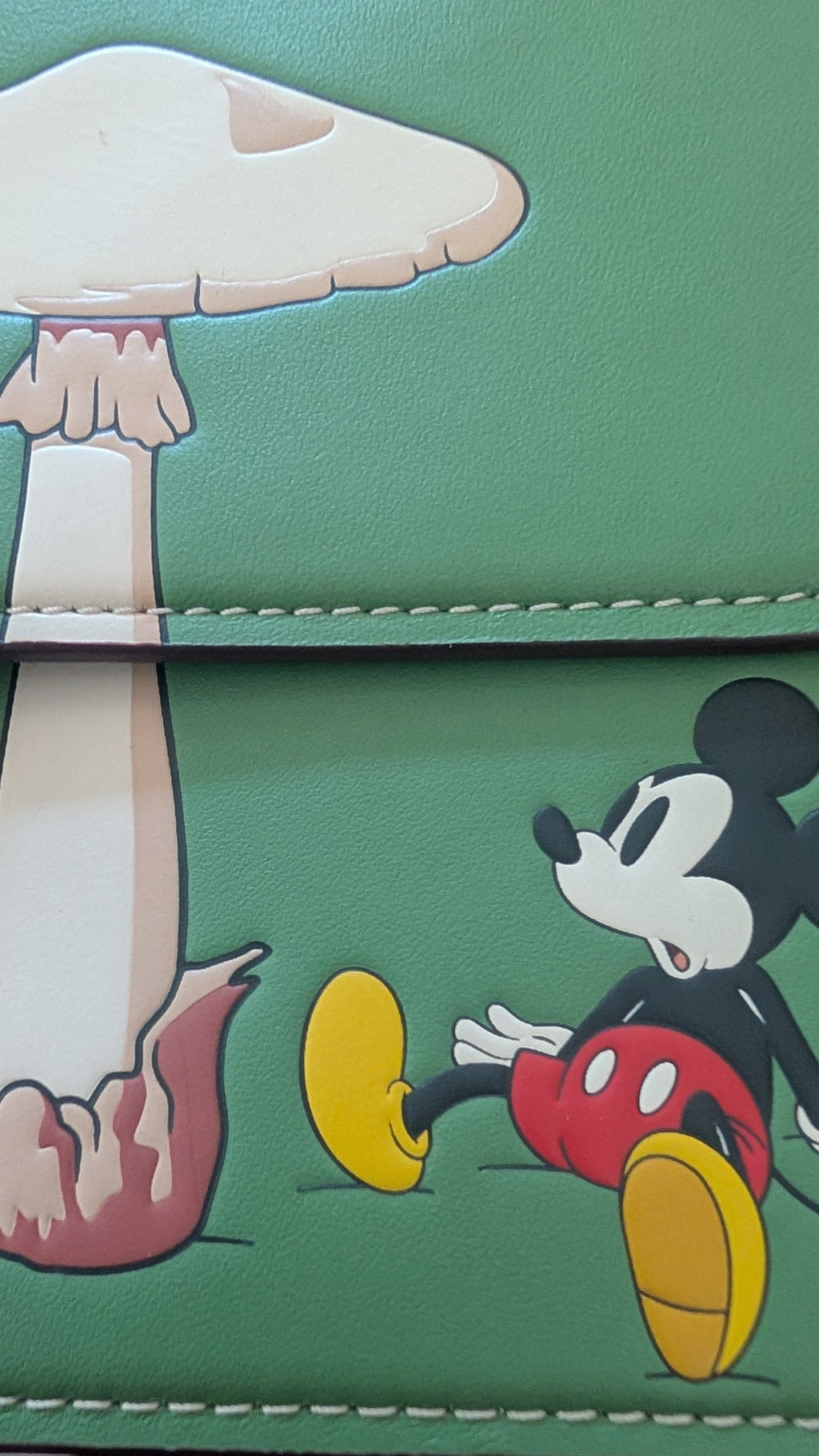 A close-up of a green faux-leather wallet featuring Mickey Mouse and Minnie Mouse designs, with a mushroom motif on the cover. Pre-Owned Coach x Disney Tabby Shoulder Bag – Mickey Mouse – Green – Limited Edition