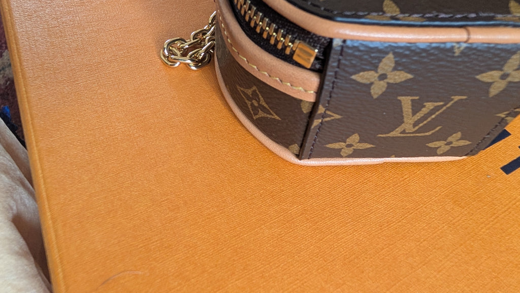 A close-up of a Louis Vuitton monogram pouch or small bag corner, showing dark brown coated canvas with tan monogram motifs, a light beige leather trim, and a gold-tone zipper pull on a textured orange surface. Pre-Owned Louis Vuitton Monogram Mini Boîte Chapeau Crossbody Bag – Full Set