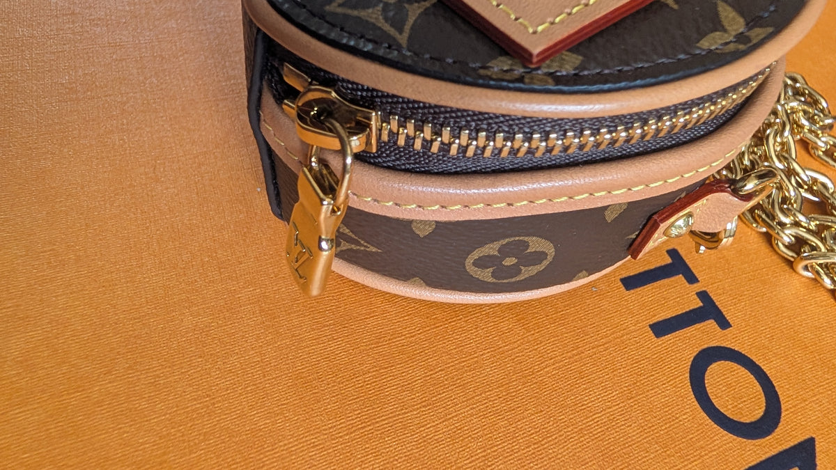 A close-up of a Louis Vuitton monogrammed cylindrical pouch or coin purse with a gold zipper, gold padlock, and gold chain strap, resting on a textured orange surface. Pre-Owned Louis Vuitton Monogram Mini Boîte Chapeau Crossbody Bag – Full Set