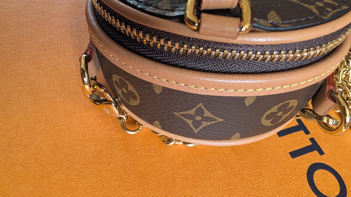 Close-up of a Louis Vuitton monogram handbag showing the brown coated canvas with gold-tone LV monogram print, tan leather trim, a gold zipper, and part of the gold chain strap on a tan textured surface. Pre-Owned Louis Vuitton Monogram Mini Boîte Chapeau Crossbody Bag – Full Set