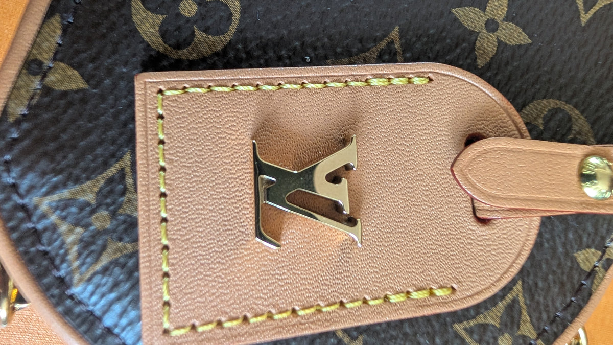 Close-up of a Louis Vuitton monogram bag featuring a tan leather luggage tag with a gold-tone LV logo clasp and yellow stitching. Pre-Owned Louis Vuitton Monogram Mini Boîte Chapeau Crossbody Bag – Full Set