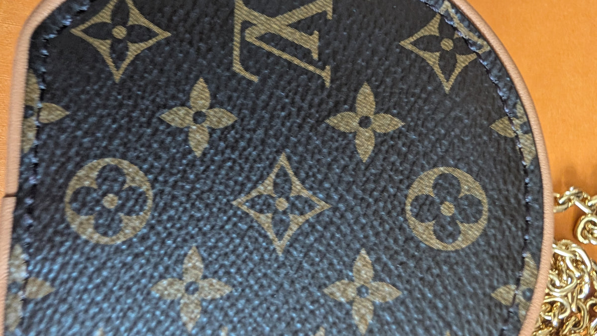 Close-up of a Louis Vuitton monogram canvas item, showing the dark brown coated canvas with tan/gold LV logos and floral motifs, plus a small portion of gold chain hardware and a tan leather edge. Pre-Owned Louis Vuitton Monogram Mini Boîte Chapeau Crossbody Bag – Full Set