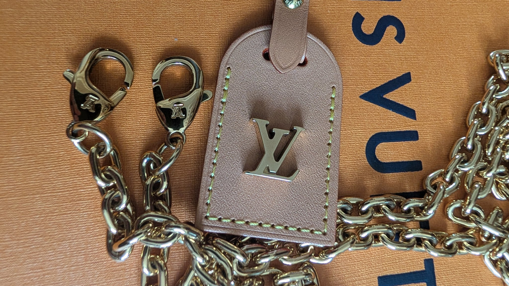 A close-up of a light beige leather luggage tag with a silver-tone metal LV monogram and a matching silver-tone chain, laid on a tan textured surface with partial blue and black lettering visible. Pre-Owned Louis Vuitton Monogram Mini Boîte Chapeau Crossbody Bag – Full Set