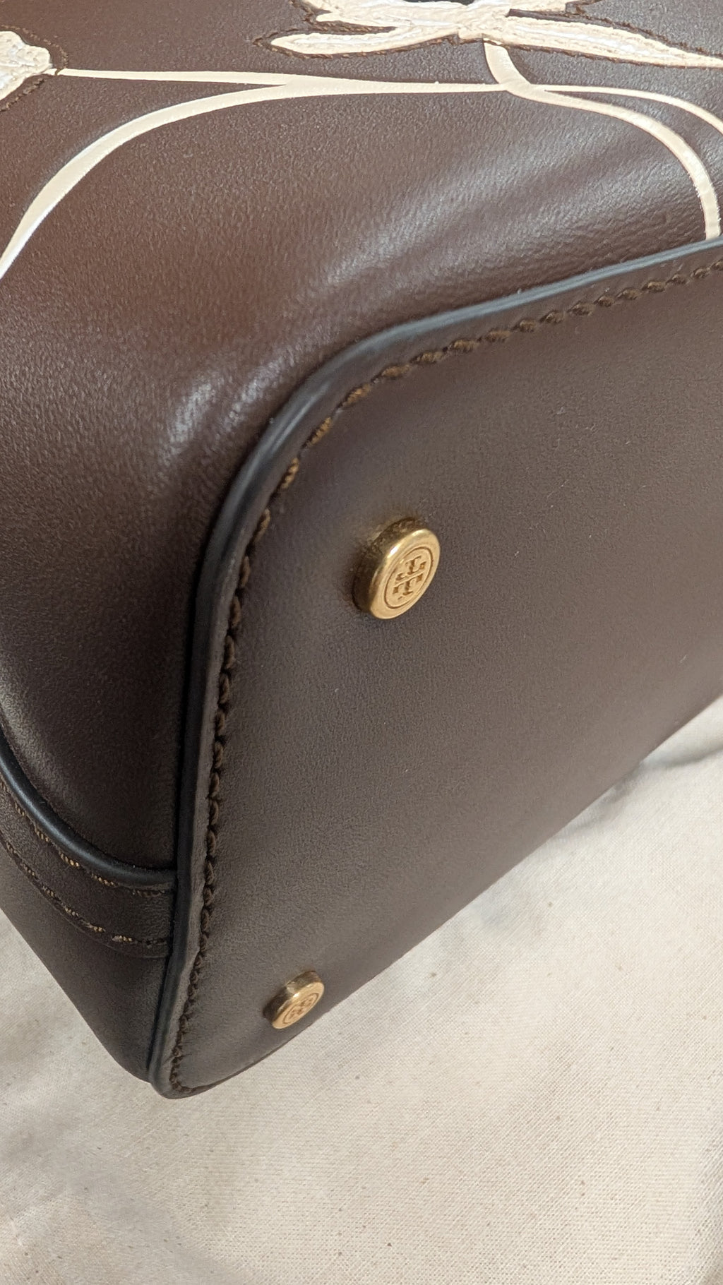 Close-up of a dark brown leather handbag corner showing gold-tone hardware and white printed line details on the upper portion. Pre-Owned Tory Burch  Floral Applique Top Handle Bag