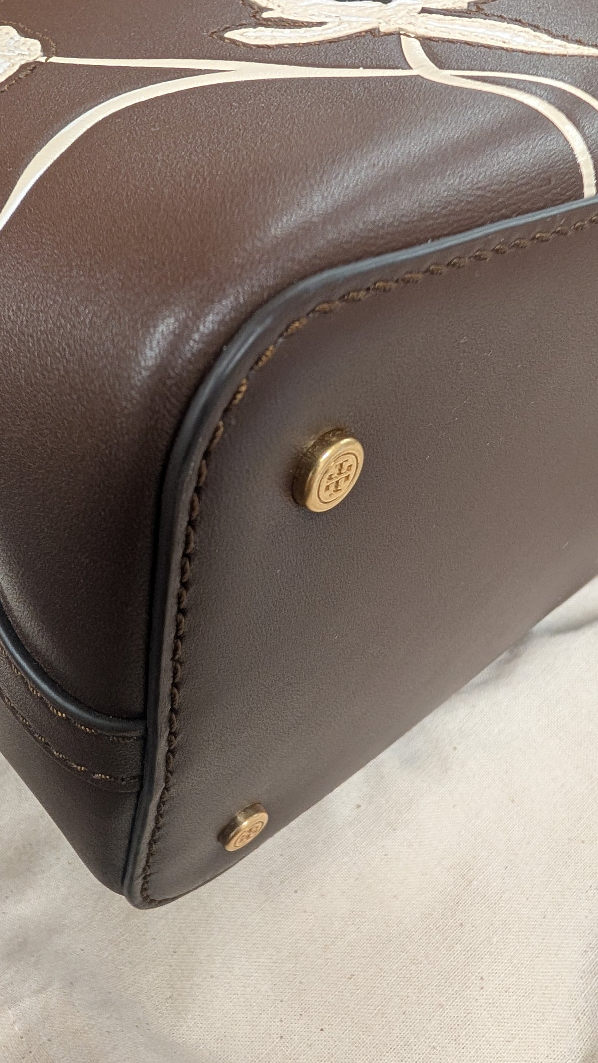 Close-up of a dark brown leather handbag corner showing gold-tone hardware and white printed line details on the upper portion. Pre-Owned Tory Burch  Floral Applique Top Handle Bag