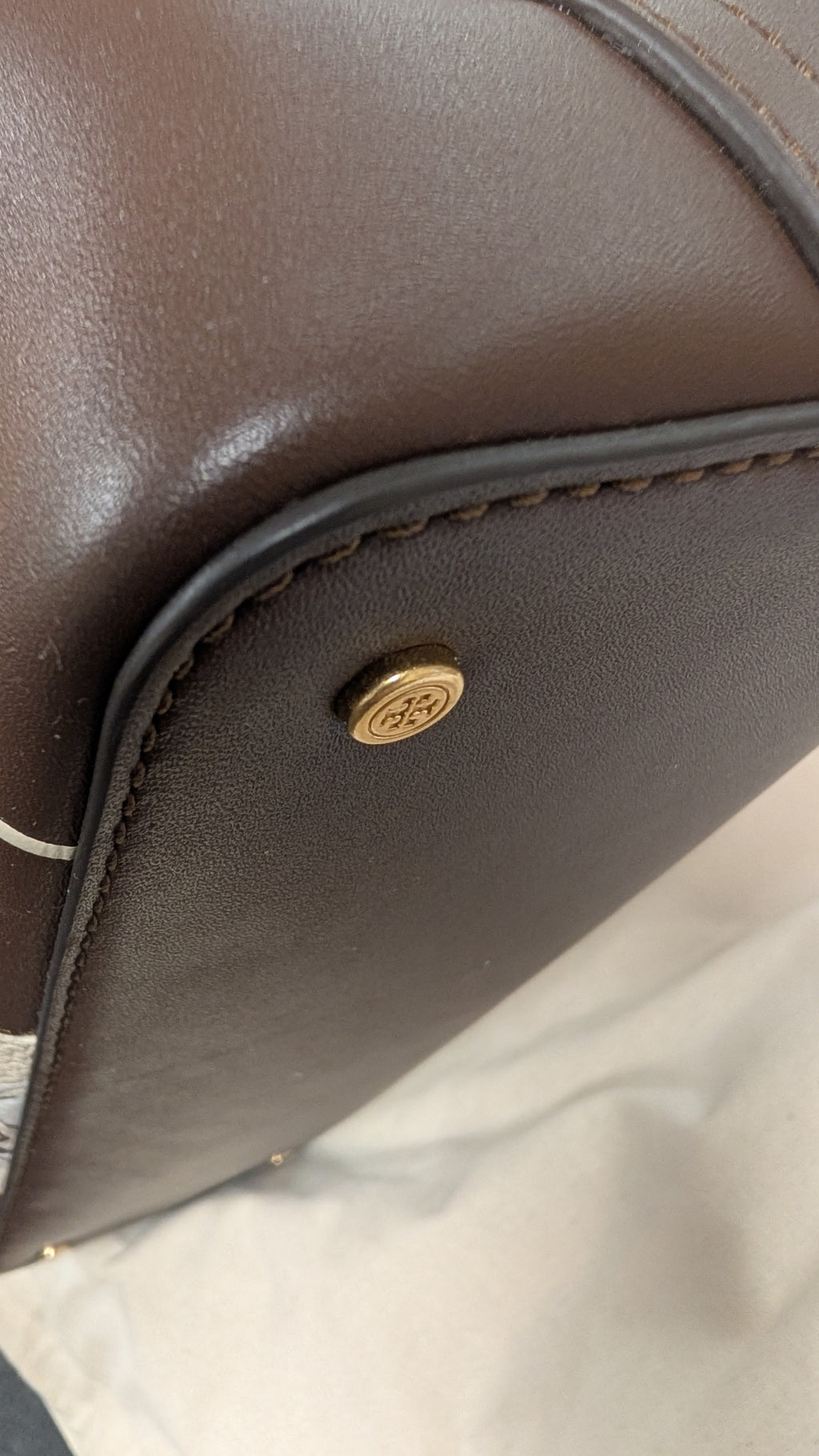 Close-up of a dark brown leather bag corner showing a gold-tone metal logo stud and stitching on a light fabric surface. Pre-Owned Tory Burch  Floral Applique Top Handle Bag