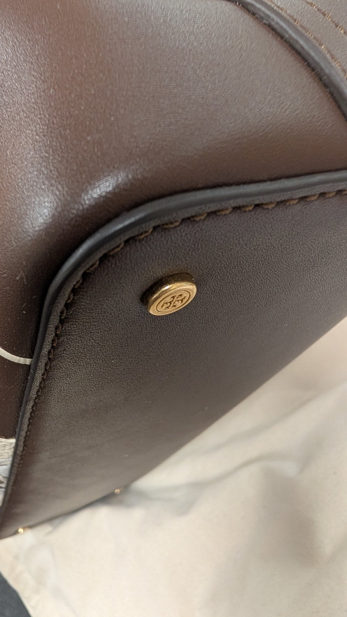 Close-up of a dark brown leather bag corner showing a gold-tone metal logo stud and stitching on a light fabric surface. Pre-Owned Tory Burch  Floral Applique Top Handle Bag