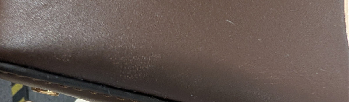 Close-up of a brown leather handbag corner with visible wear, scuffs, and dirt on the surface, resting on a white fabric surface. Pre-Owned Tory Burch  Floral Applique Top Handle Bag