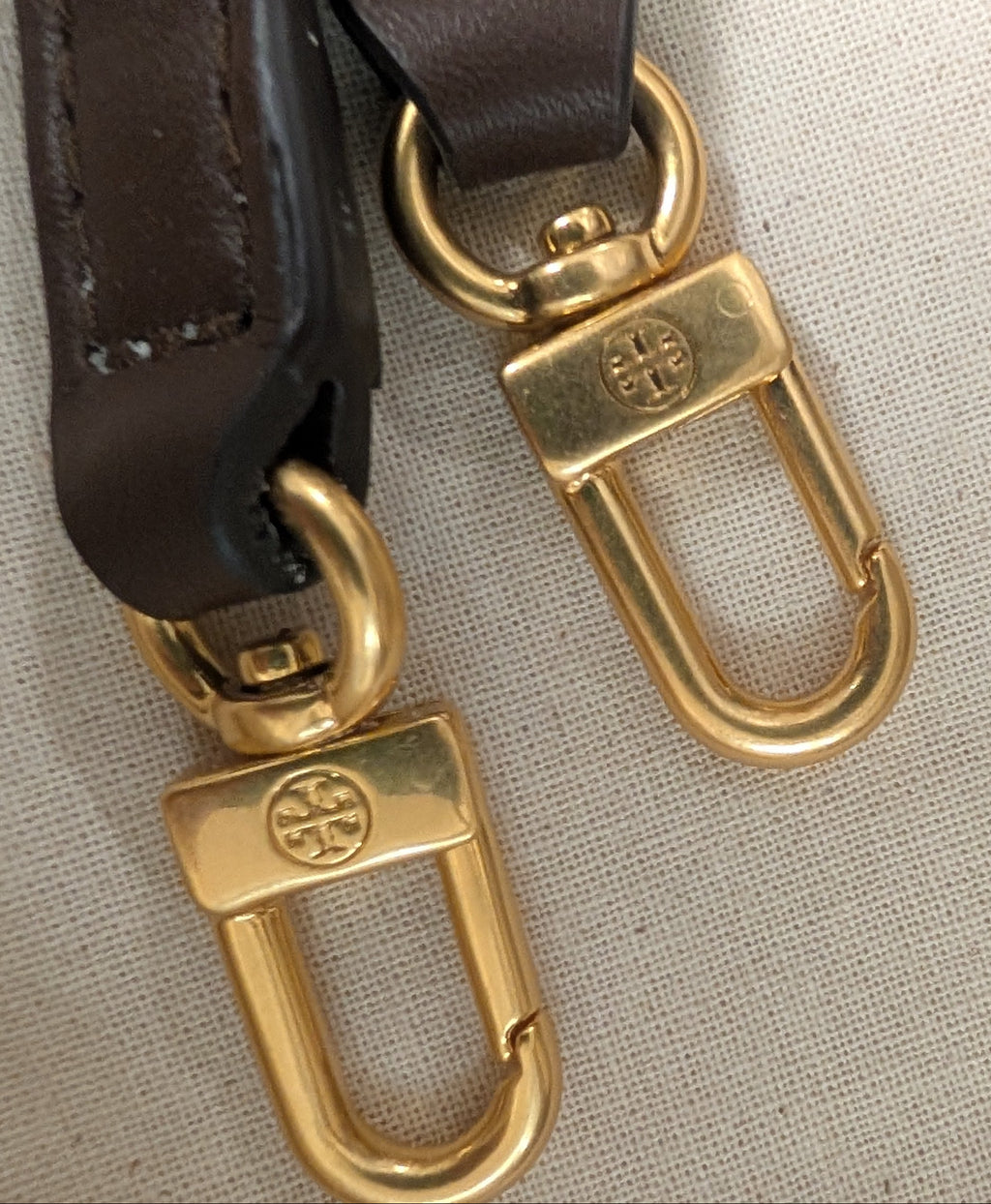 Close-up of a dark brown leather strap with gold-tone metal hardware, including two lobster clasp rings, displayed on a textured beige fabric surface. Pre-Owned Tory Burch  Floral Applique Top Handle Bag