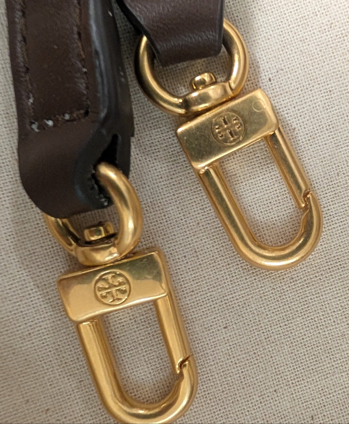 Close-up of a dark brown leather strap with gold-tone metal hardware, including two lobster clasp rings, displayed on a textured beige fabric surface. Pre-Owned Tory Burch  Floral Applique Top Handle Bag
