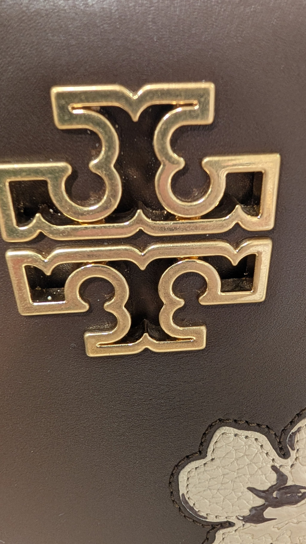 Close-up of a dark brown leather bag featuring a prominent gold-tone Tory Burch logo emblem and a small portion of a white embroidered monogram. Pre-Owned Tory Burch  Floral Applique Top Handle Bag