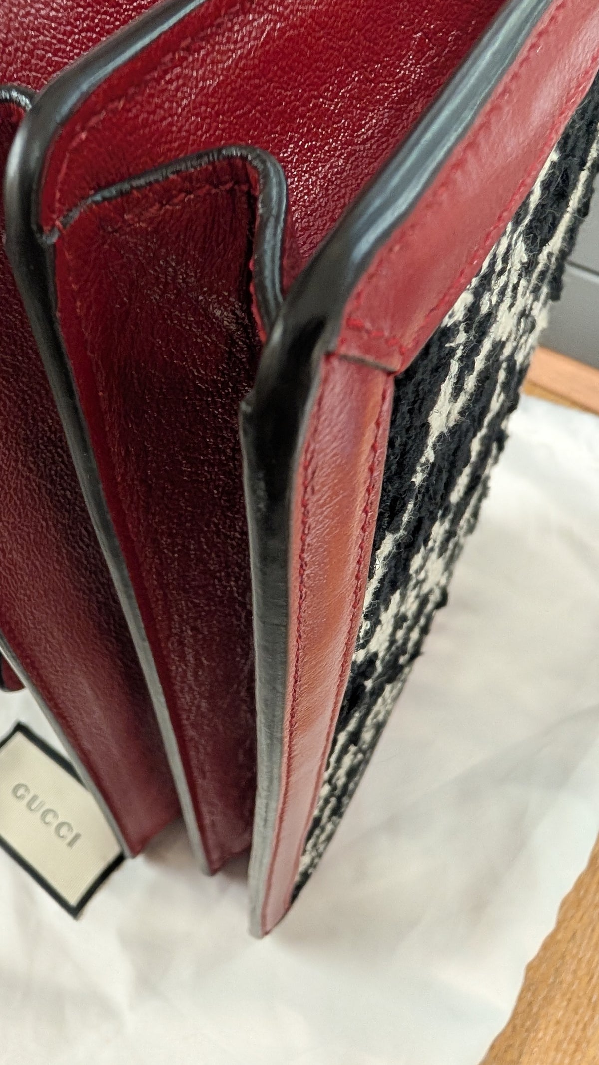 Close-up of a Gucci leather bag corner showing deep red leather trim, black edging, a black-and-white patterned textile side, and a small Gucci tag on white dust bag paper. Pre-Owned Gucci Dionysus GG Tweed Medium Shoulder Bag – Red/Black