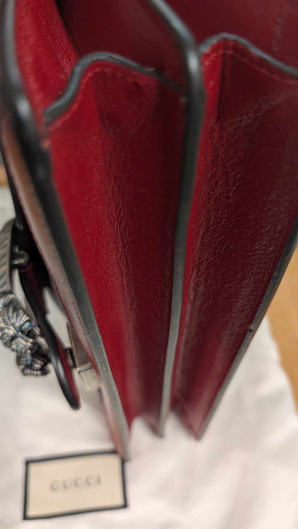 Close-up of a red leather Gucci bag with dark trim and a silver-tone chain, resting on a white Gucci dust bag with a Gucci label visible. Pre-Owned Gucci Dionysus GG Tweed Medium Shoulder Bag – Red/Black