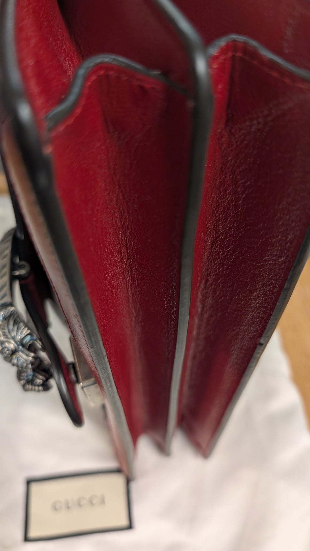 Close-up of a red leather Gucci bag with dark trim and a silver-tone chain, resting on a white Gucci dust bag with a Gucci label visible. Pre-Owned Gucci Dionysus GG Tweed Medium Shoulder Bag – Red/Black