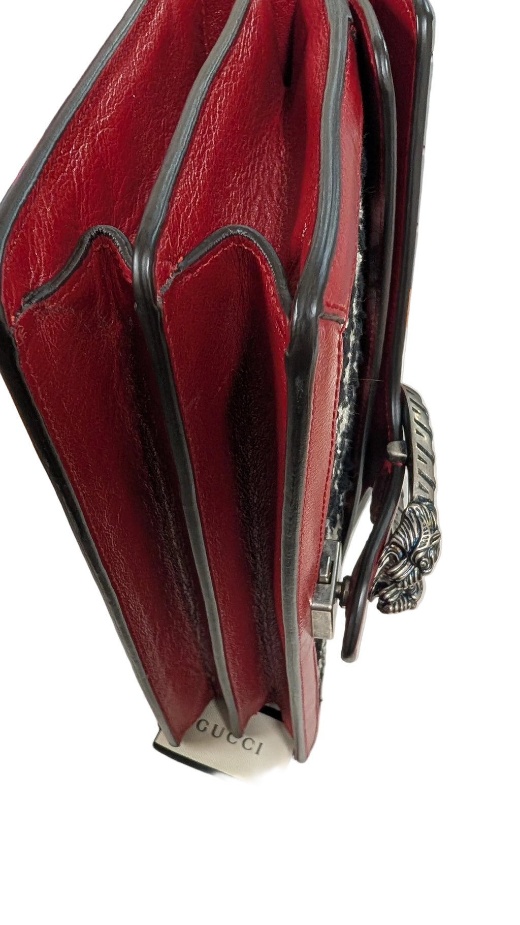 A red Gucci leather bag shown from a side angle, standing on a white surface with a Gucci-branded card visible at the base. The bag has dark trim and a patterned strap detail on the right side. Pre-Owned Gucci Dionysus GG Tweed Medium Shoulder Bag – Red/Black