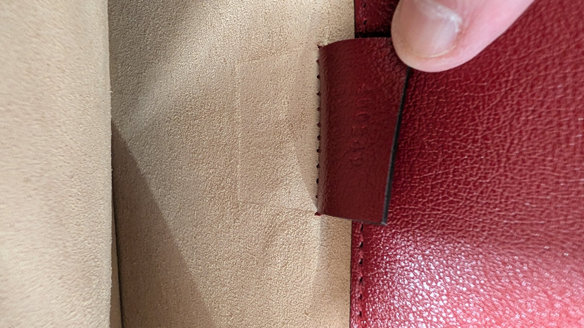 Close-up of a red leather item being held by a finger against a beige suede-like background, showing a small embossed brand mark on the red tab. Pre-Owned Gucci Dionysus GG Tweed Medium Shoulder Bag – Red/Black
