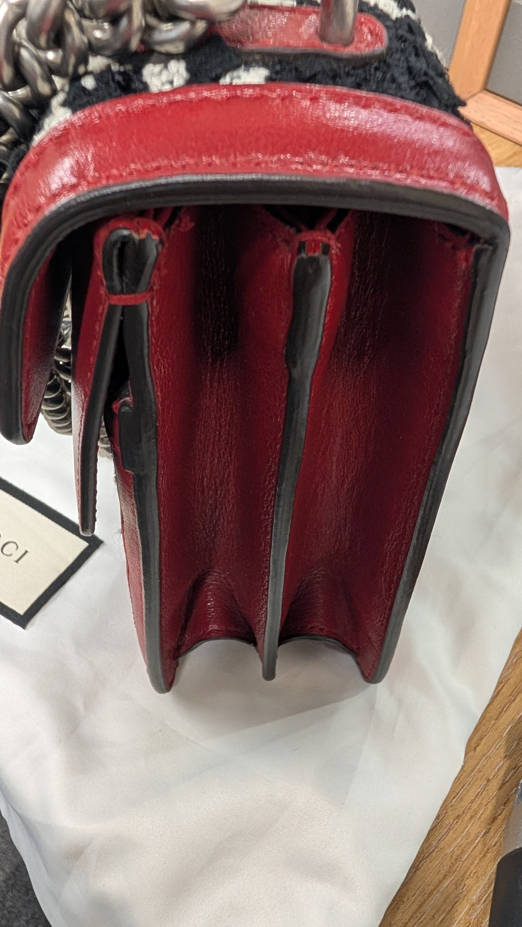 Close-up of a red leather handbag showing the interior side with black trim, a black-and-white patterned textile detail, and part of a chain strap on a white fabric surface. Pre-Owned Gucci Dionysus GG Tweed Medium Shoulder Bag – Red/Black