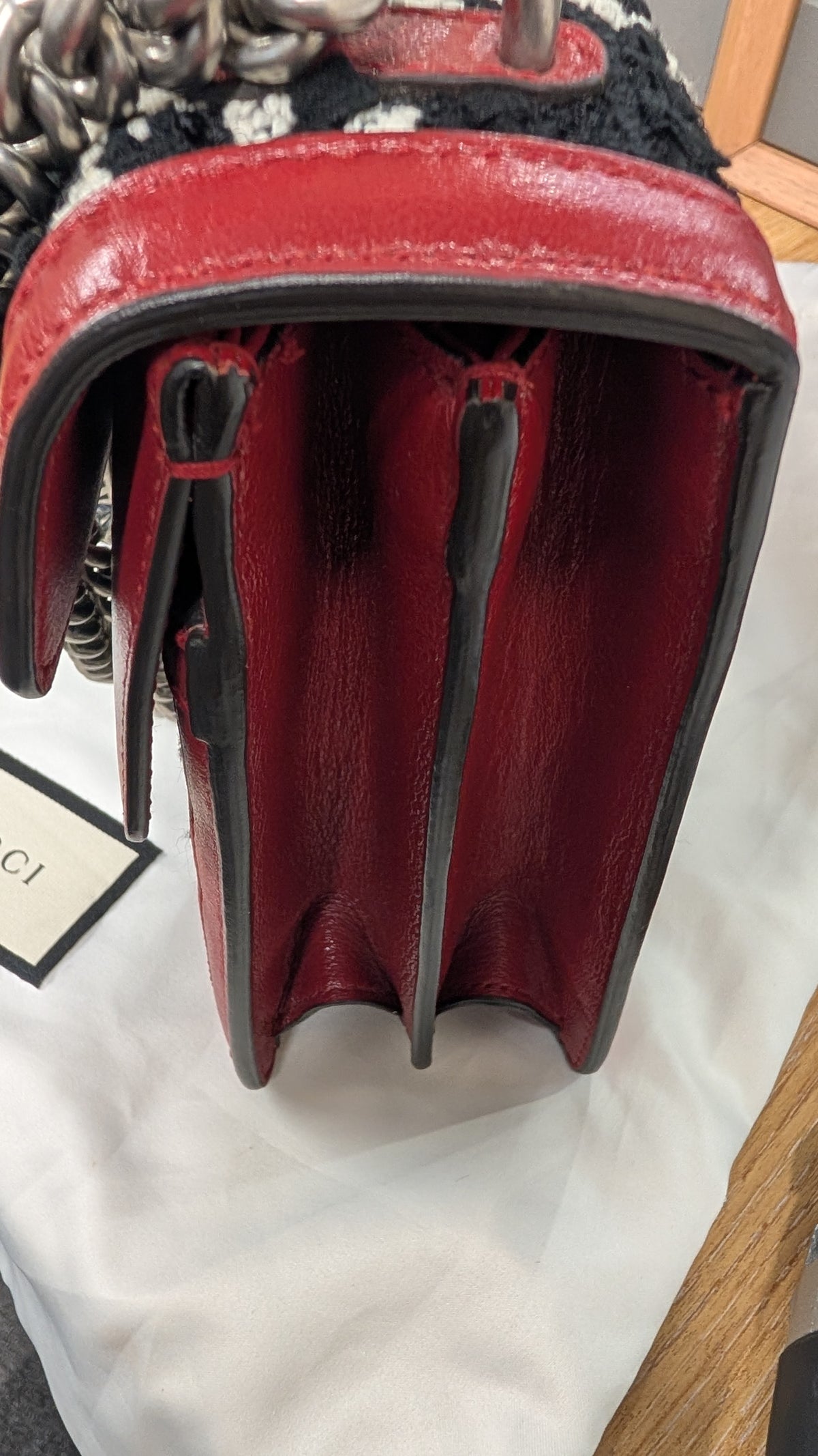 Close-up of a red leather handbag showing the interior side with black trim, a black-and-white patterned textile detail, and part of a chain strap on a white fabric surface. Pre-Owned Gucci Dionysus GG Tweed Medium Shoulder Bag – Red/Black