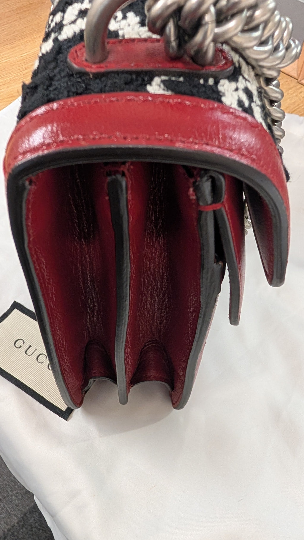 Close-up of a Gucci handbag showing the interior lining, chain strap, and textured black-and-white fabric exterior, resting on a white dust bag. Pre-Owned Gucci Dionysus GG Tweed Medium Shoulder Bag – Red/Black