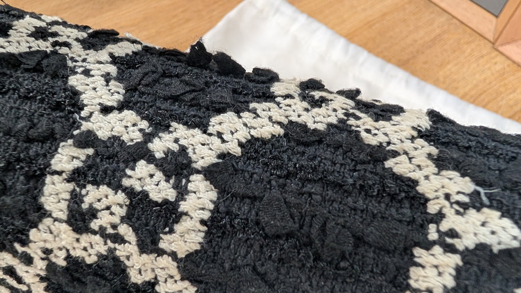 A close-up of a black and off-white textured rug or cushion cover with a chunky, looped fabric pattern, shown on a wooden floor next to a white fabric piece. Pre-Owned Gucci Dionysus GG Tweed Medium Shoulder Bag – Red/Black