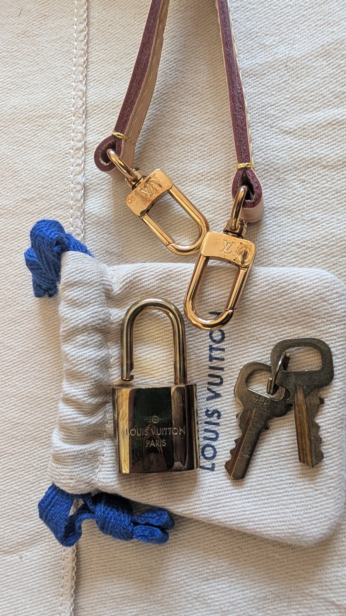 Louis Vuitton padlock keychain with gold-toned padlock, brown leather strap, and two keys, displayed on a white drawstring pouch with Louis Vuitton branding. Pre-Owned Louis Vuitton Alma BB Monogram World Tour / Travel Sticker Shoulder Bag