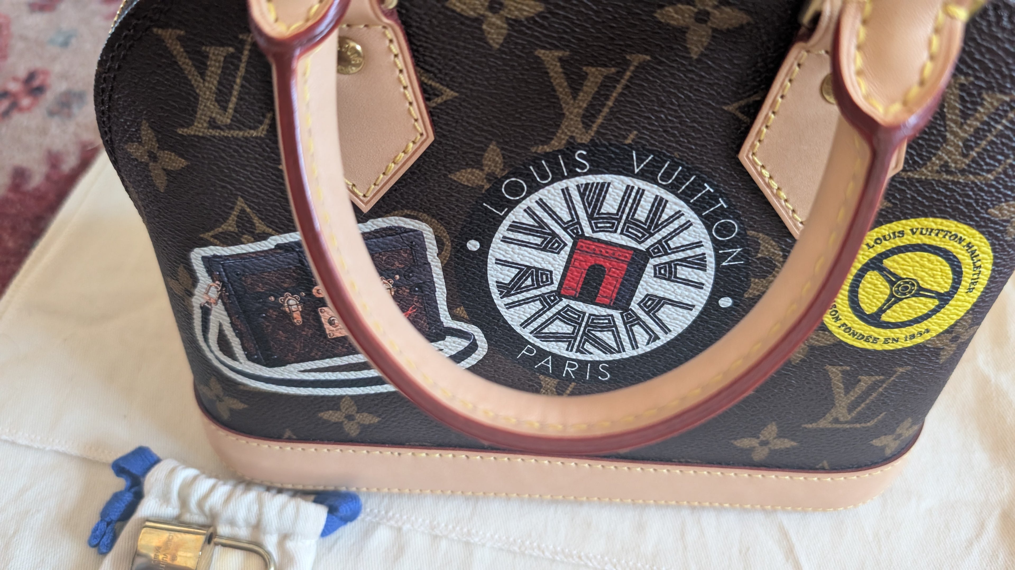A Louis Vuitton handbag in brown monogram canvas with beige leather trim and handles, featuring a circular Louis Vuitton Paris patch and a yellow circular patch on the front, photographed on a light fabric surface. Pre-Owned Louis Vuitton Alma BB Monogram World Tour / Travel Sticker Shoulder Bag