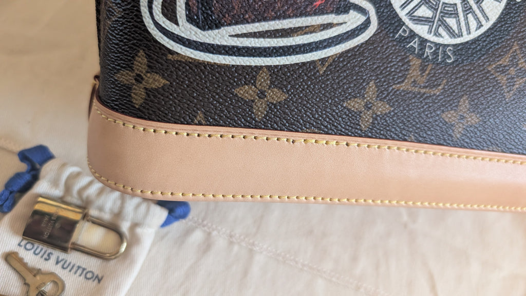 Close-up of a Louis Vuitton monogram handbag corner showing dark brown coated canvas with tan monogram flowers, a beige leather trim, and part of the white Paris logo. A Louis Vuitton dust bag and small lock are visible on a light fabric surface. Pre-Owned Louis Vuitton Alma BB Monogram World Tour / Travel Sticker Shoulder Bag