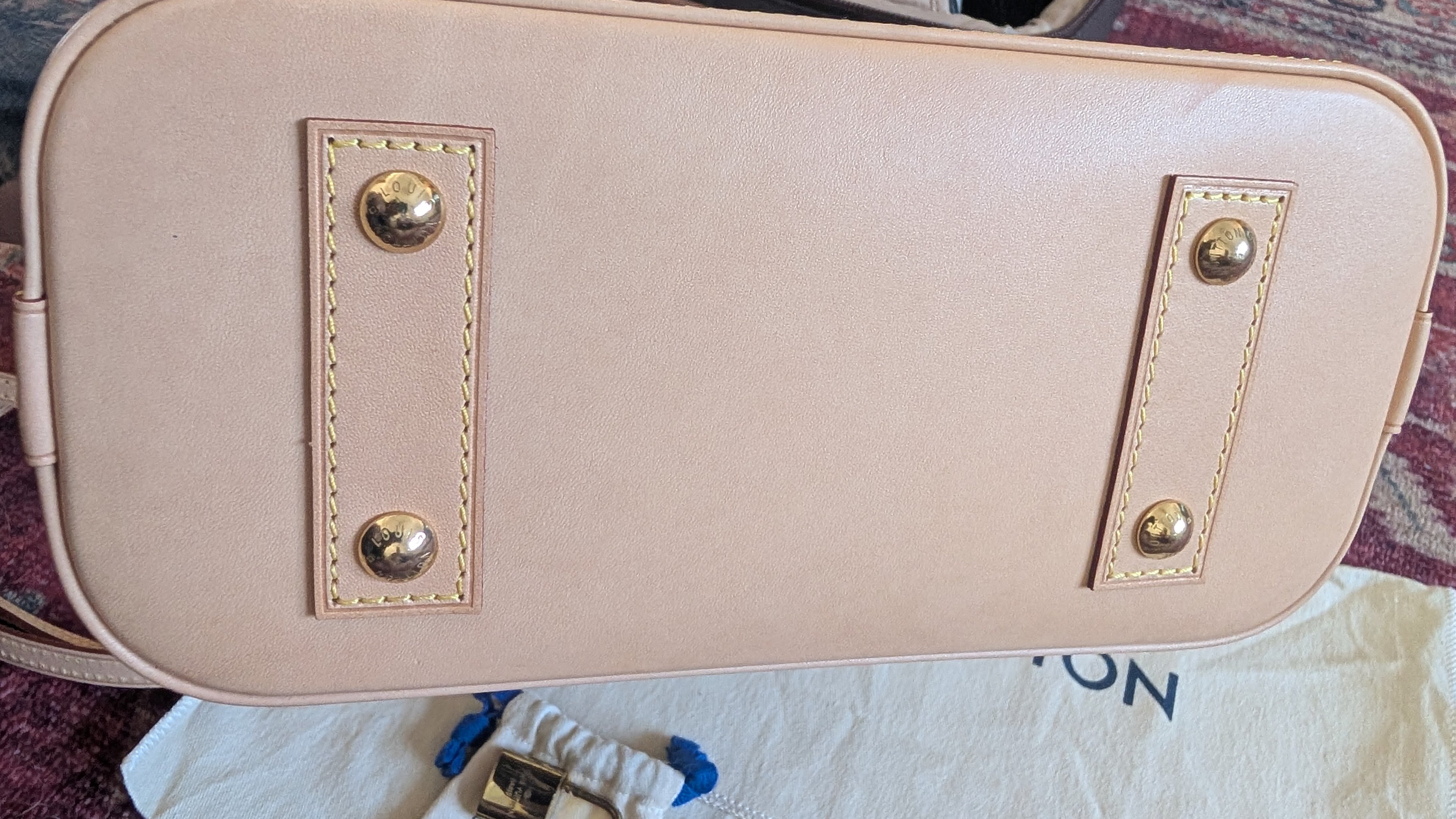 A close-up of a light blush pink leather handbag bottom, showing gold-tone metal studs and rivets on a white dust bag. Pre-Owned Louis Vuitton Alma BB Monogram World Tour / Travel Sticker Shoulder Bag