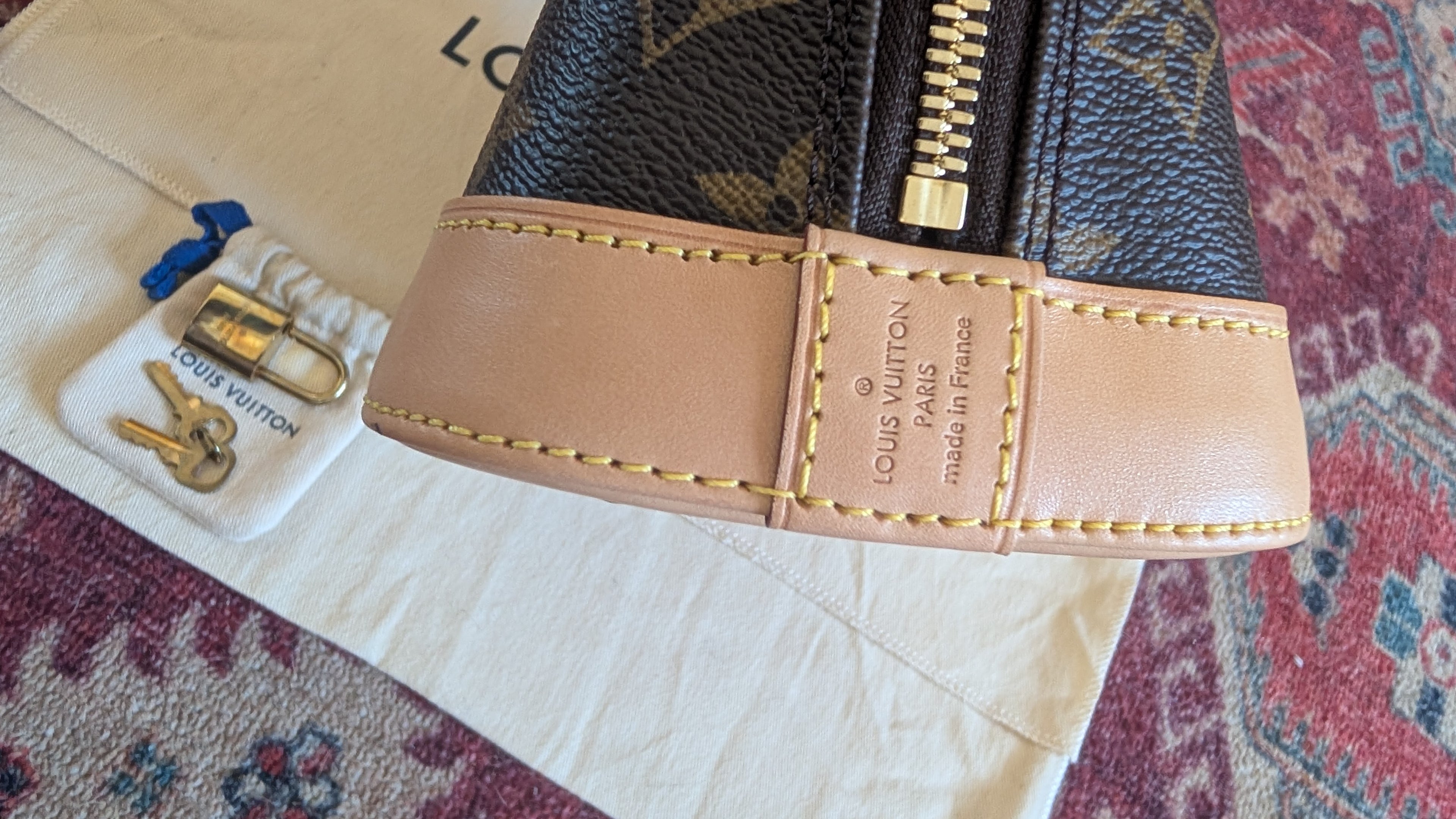 Close-up of a Louis Vuitton monogram leather bag with a tan leather trim and gold zipper, placed on a cream dust bag with a small branded tag and keys on a patterned rug. Pre-Owned Louis Vuitton Alma BB Monogram World Tour / Travel Sticker Shoulder Bag