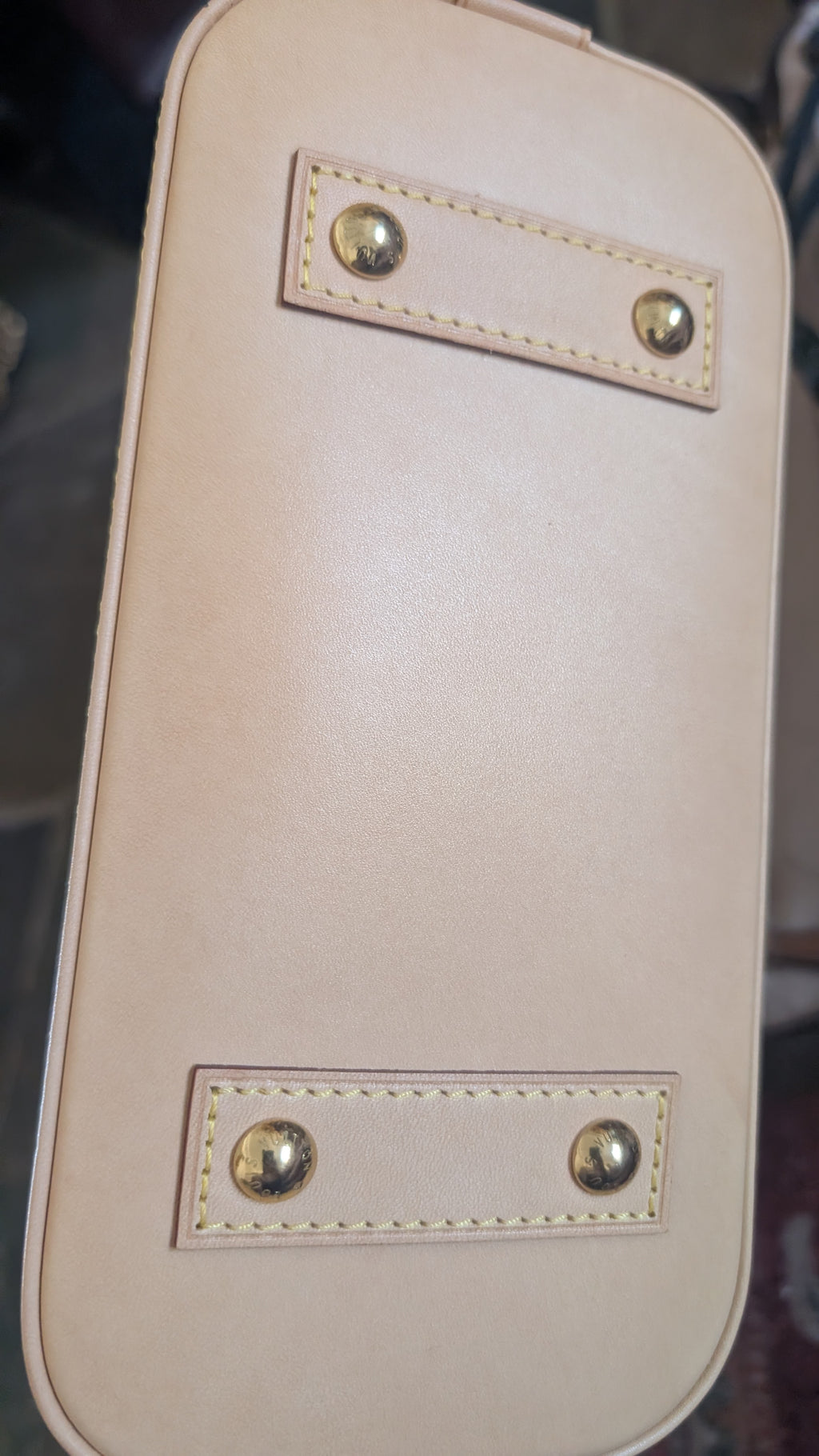 Close-up of the bottom corner of a light beige leather bag, showing two rectangular stitched leather tabs with gold-tone metal rivets. Pre-Owned Louis Vuitton Alma BB Monogram World Tour / Travel Sticker Shoulder Bag