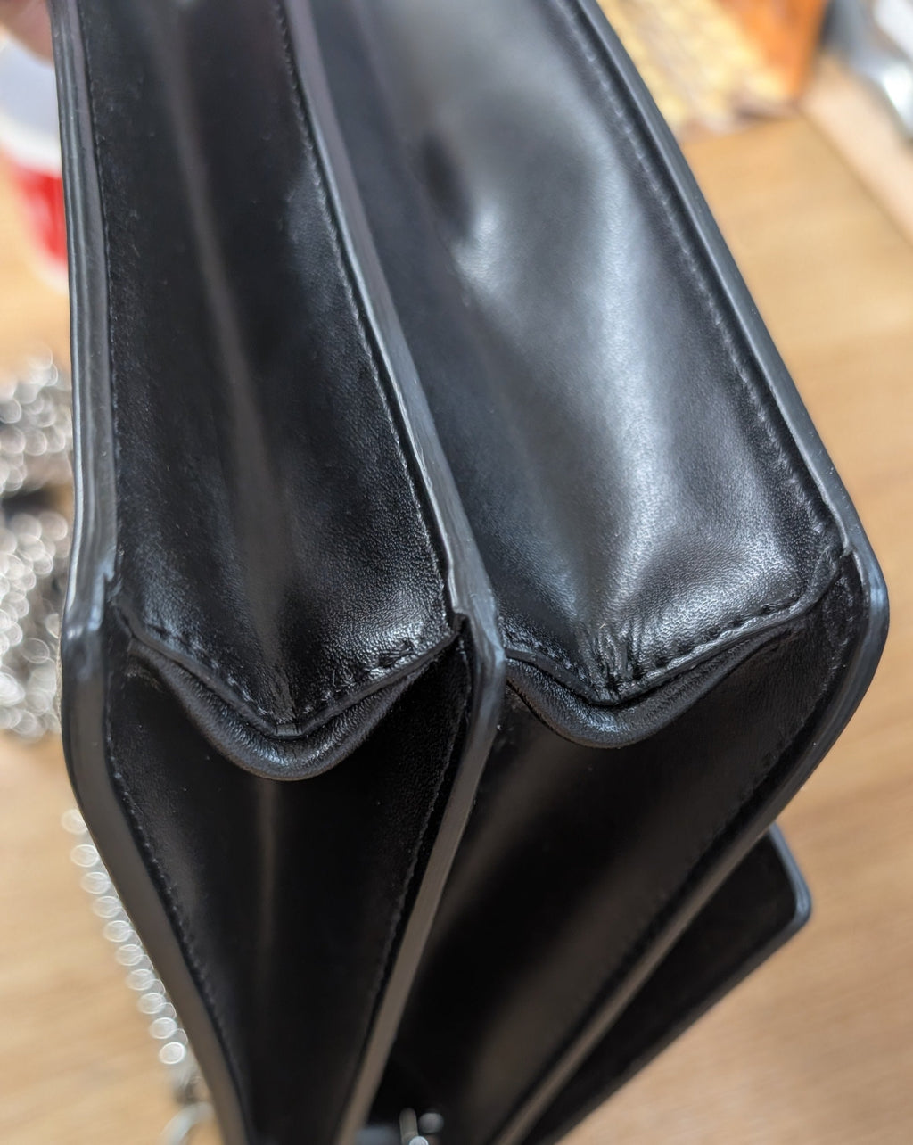 Close-up of a black leather handbag corner with a silver chain strap, resting on a light wood surface. Pre-Owned Fendi Kan I Monster Eyes Black Leather Chain Shoulder Bag
