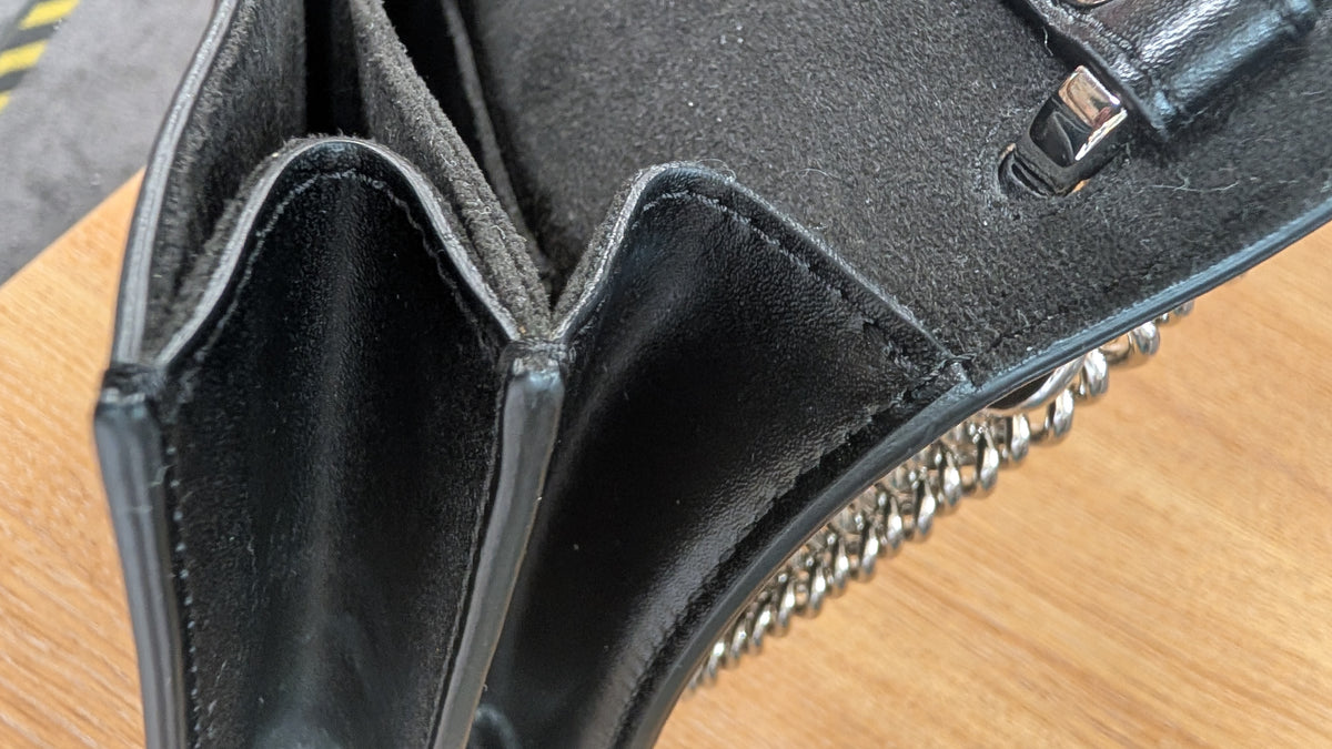 Close-up detail of a black leather handbag showing the interior lining, fold-over flap, silver-tone chain detail, and a portion of the exterior strap on a wooden surface. Pre-Owned Fendi Kan I Monster Eyes Black Leather Chain Shoulder Bag
