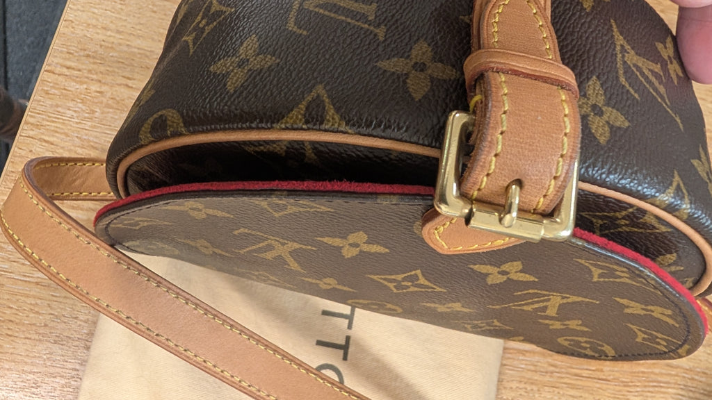 A close-up of a Louis Vuitton monogram handbag in brown coated canvas with tan leather trim and a gold-tone buckle, resting on a light wood surface with a dust bag partially visible underneath. Pre-Owned Louis Vuitton Monogram Tambourin Shoulder / Crossbody Bag