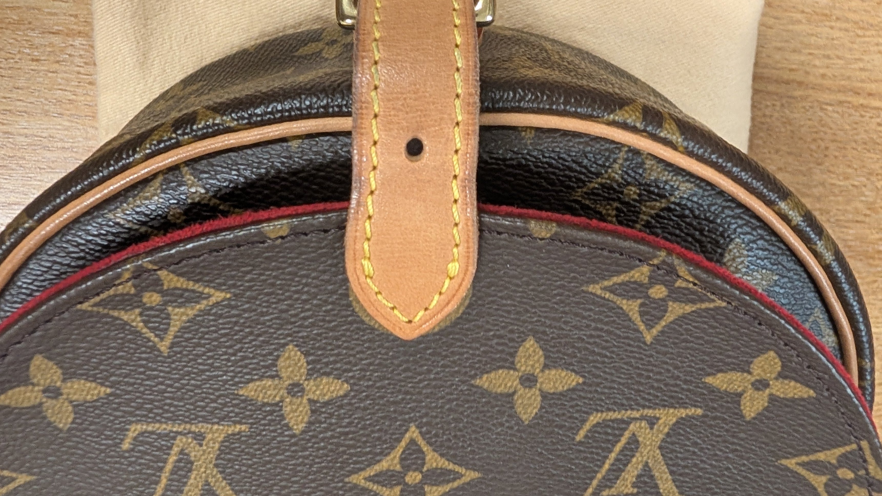 Close-up of a Louis Vuitton monogram bag in brown coated canvas with tan leather trim and a red piping accent, resting on a light wood surface. Pre-Owned Louis Vuitton Monogram Tambourin Shoulder / Crossbody Bag