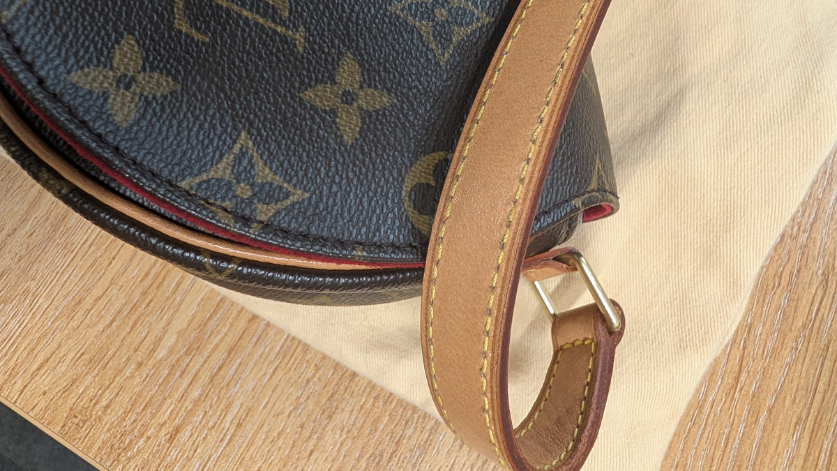 Close-up of a Louis Vuitton monogram bag in dark brown coated canvas with a tan leather strap, resting on a light beige fabric surface over a wooden table. Pre-Owned Louis Vuitton Monogram Tambourin Shoulder / Crossbody Bag