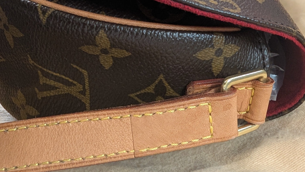 A close-up of a Louis Vuitton monogram canvas shoulder bag with a tan leather strap and gold-tone hardware, resting on a beige dust bag over a wooden surface. Pre-Owned Louis Vuitton Monogram Tambourin Shoulder / Crossbody Bag