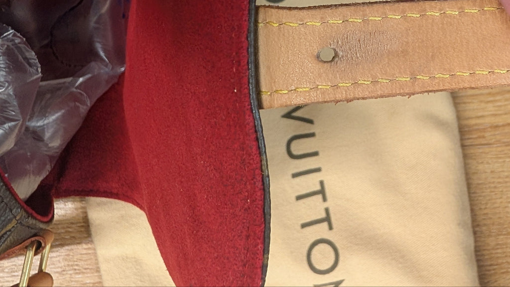 A close-up of a Louis Vuitton canvas dust bag with a tan leather strap, held by a hand. The red velvet-like fabric of the bag is visible on the left, and the beige canvas print with black 'LOUIS VUITTON' lettering is in the background on a wooden surface. Pre-Owned Louis Vuitton Monogram Tambourin Shoulder / Crossbody Bag