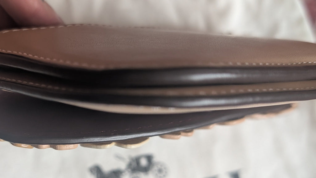 A close-up of a person's hand holding a brown leather Coach wallet or card holder over a white Coach dust bag with the Coach logo visible. Pre-Owned Coach Courier Wristlet with Floral Appliqué – Glovetanned Leather