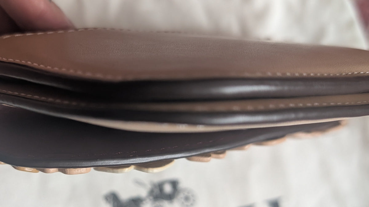 A close-up of a person's hand holding a brown leather Coach wallet or card holder over a white Coach dust bag with the Coach logo visible. Pre-Owned Coach Courier Wristlet with Floral Appliqué – Glovetanned Leather